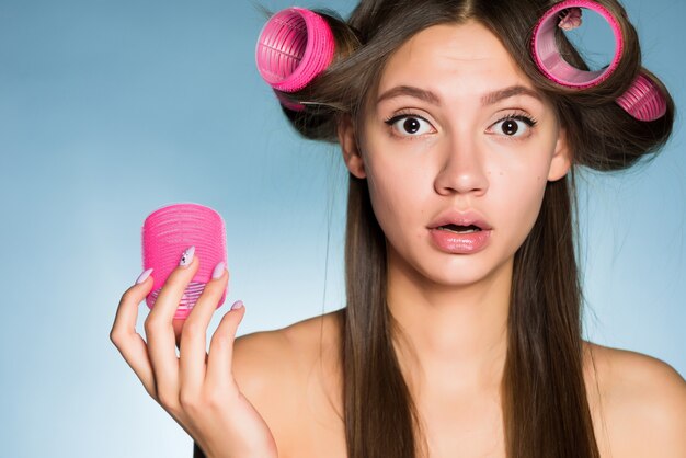 Surprised young girl wants a beautiful hairstyle, on her head big pink hair curlers