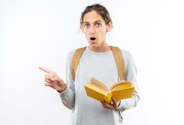 Surprised young guy student wearing backpack holding book points at side 