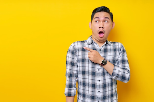 Surprised young handsome Asian man 20s wearing a plaid shirt pointing a finger at the advertising area isolated on yellow background People lifestyle concept