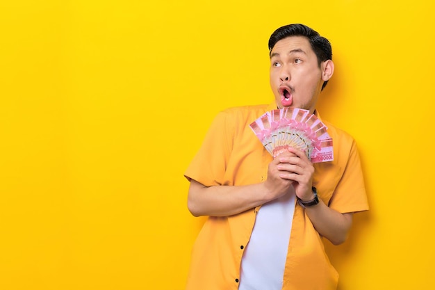 Surprised young handsome asian man holding bunch of money and looking away at copy space isolated on yellow background