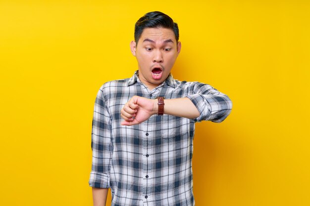 Surprised young handsome asian man wearing a white checkered shirt looking at his watch to check time isolated over yellow background people lifestyle concept