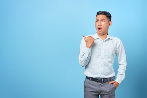 Surprised young handsome businessman pointing finger away at copy space on blue background