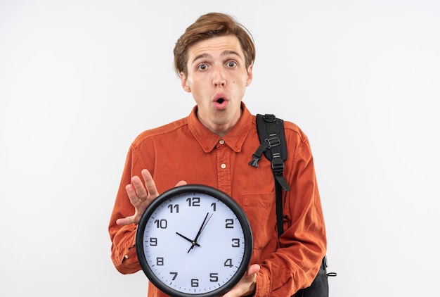 Sorpreso giovane bel ragazzo che indossa una camicia rossa con lo zaino che tiene l'orologio da parete isolato sul muro bianco