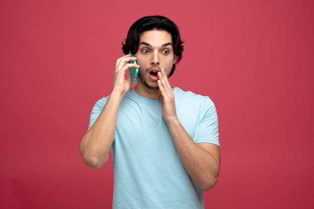 surprised young handsome man keeping hand near mouth looking at side talking on phone isolated on red background