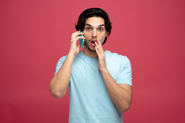 surprised young handsome man looking at camera keeping hand near mouth talking on phone isolated on red background