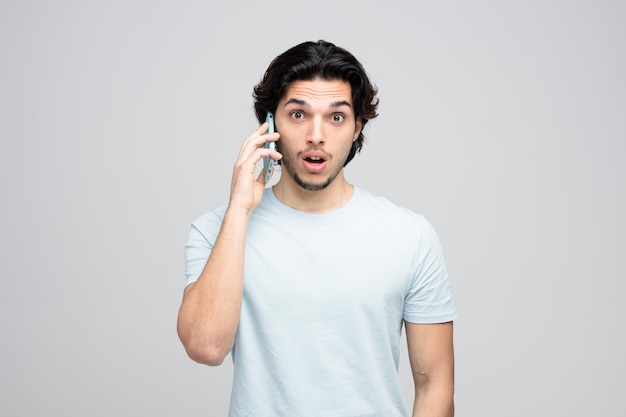 surprised young handsome man looking at camera while talking on phone isolated on white background