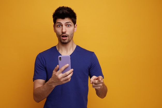 Surprised young handsome man looking and pointing at camera stretching mobile phone out towards camera on yellow background with copy space