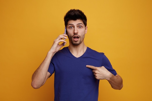 Surprised young handsome man talking on phone looking at camera pointing at himself on yellow background