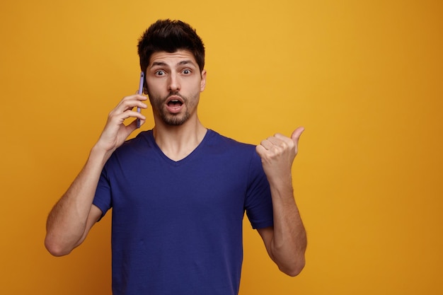 Surprised young handsome man talking on phone looking at camera pointing to side on yellow background with copy space