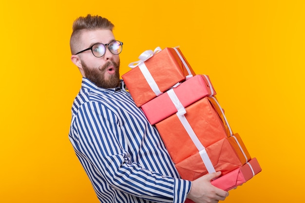 Surprised young hipster guy in glasses holding a large stack of gift boxes on a yellow wall. The concept of New Year and Christmas gifts.