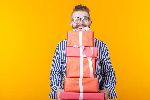 Surprised young hipster guy in glasses holding a large stack of gift boxes on a yellow wall