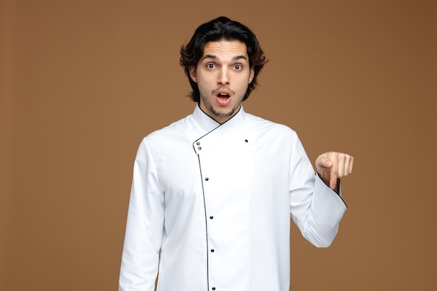 surprised young male chef wearing uniform looking at camera pointing down isolated on brown background