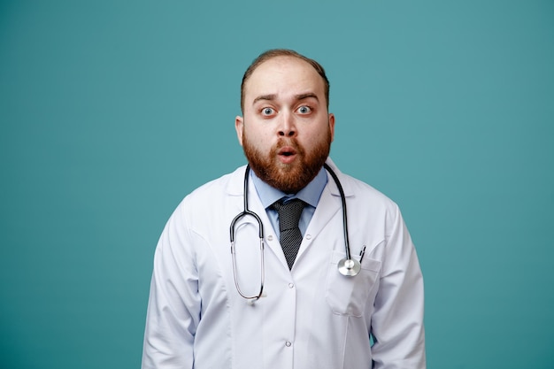 Sorpreso giovane medico maschio che indossa cappotto medico e stetoscopio intorno al collo guardando la fotocamera isolata su sfondo blu