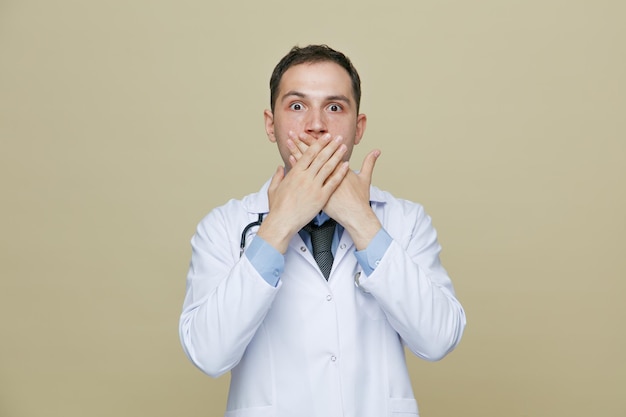 surprised young male doctor wearing medical robe and stethoscope around neck looking at camera while keeping hands on mouth isolated on olive green background