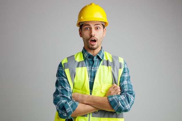 Surprised young male engineer wearing safety helmet and uniform keeping arms crossed looking at camera isolated on white background