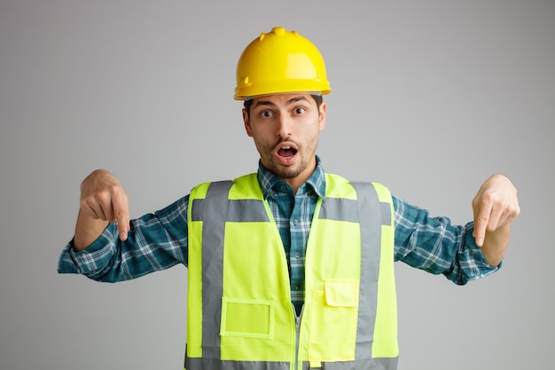 Surprised young male engineer wearing safety helmet and uniform looking at camera pointing down isolated on white background