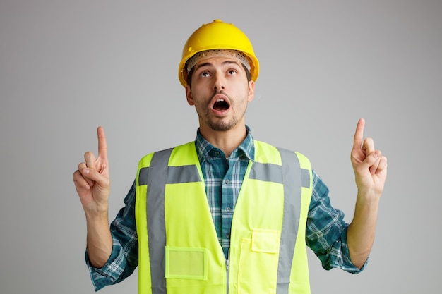 Surprised young male engineer wearing safety helmet and uniform looking up pointing fingers up isolated on white background