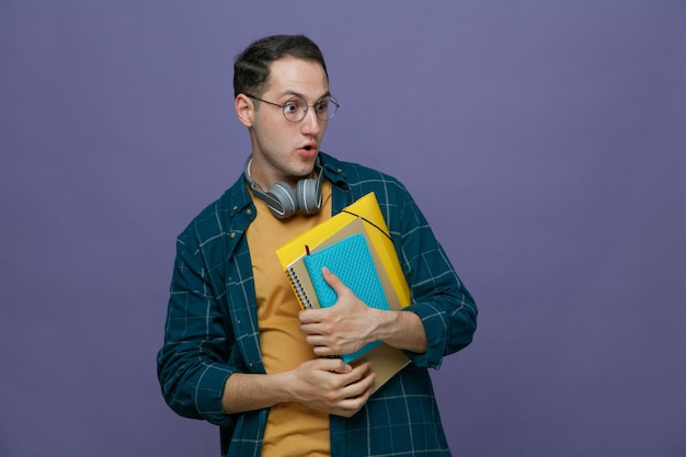 Surprised young male student wearing glasses headphones around neck holding note book note pad folder with both hands looking at side isolated on purple background