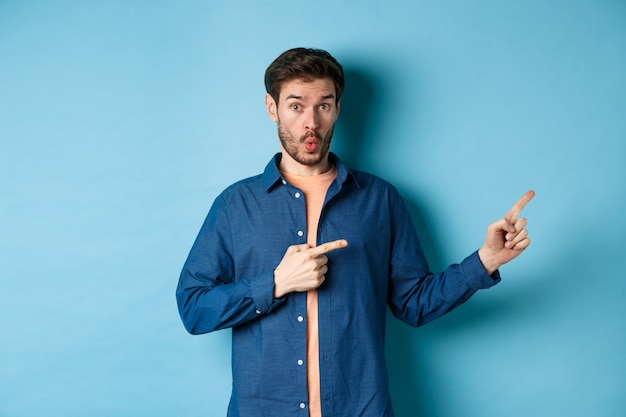 Surprised young man checking out promo, pointing fingers right at logo and saying wow, standing on blue background.