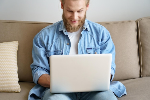 Photo surprised young man computer user winning online sport betting using laptop sitting on sofa at home office smiling man amazed by internet bid victory receiving great email exam results concept