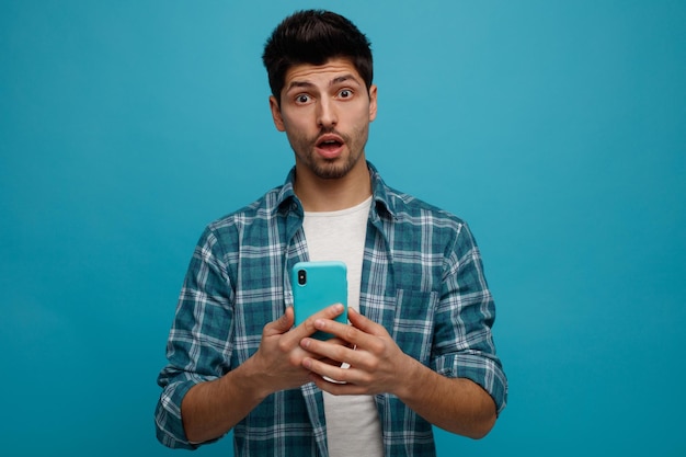 Surprised young man holding mobile phone looking at camera isolated on blue background