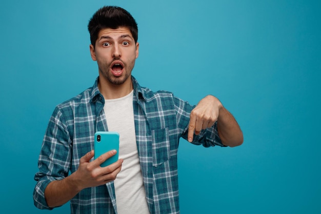 Surprised young man holding mobile phone looking at camera pointing down isolated on blue background