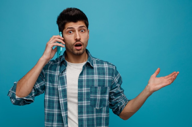 Surprised young man looking at camera showing empty hand while talking on phone isolated on blue background