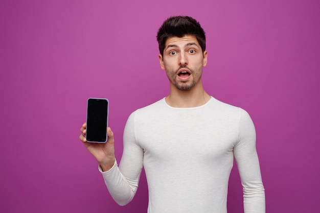 Surprised young man looking at camera showing mobile phone to camera isolated on purple background