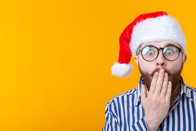 Surprised young man Santa Claus startledly closes his mouth on a yellow wall. The concept of Christmas .