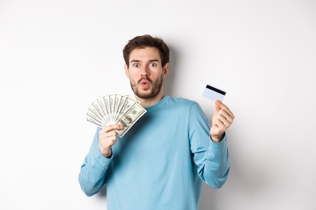 Surprised young man showing money in dollars and plastic credit card, gasping and saying wow with amazed face, standing on white background