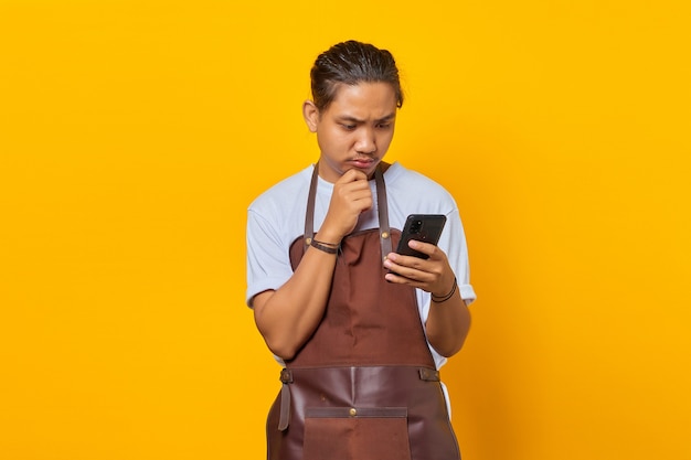 surprised young man wearing apron looking at incoming message on mobile phone with hand on chin