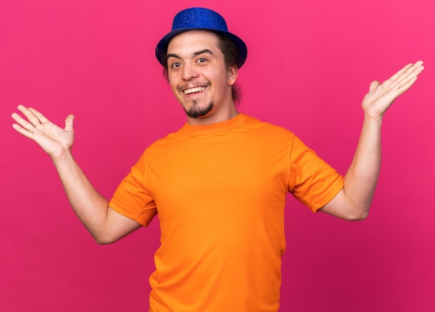 Surprised young man wearing party hat spreading hands isolated on pink wall