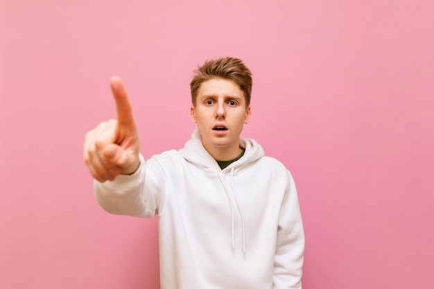 Удивленный молодой человек в белой толстовке стоит на розовом фоне и прикасается пальцем к камере