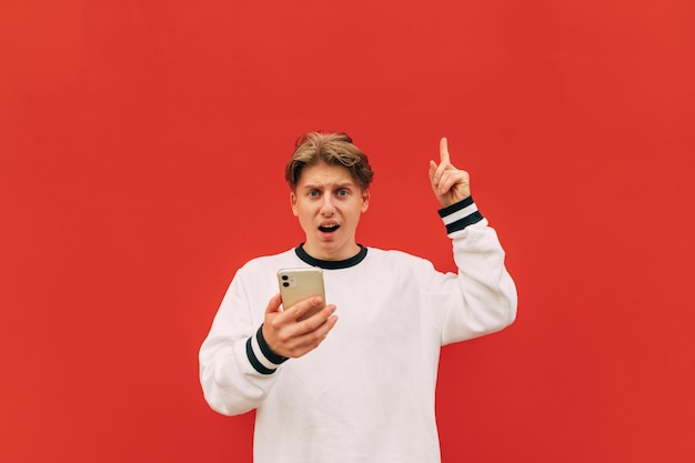 Surprised young man in white sweatshirt enjoys smartphone on red background looks at camera