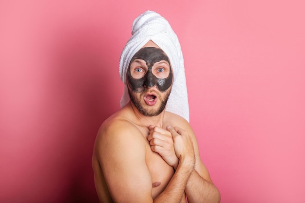 Surprised young man with cosmetic mask nude hides behind on pink background