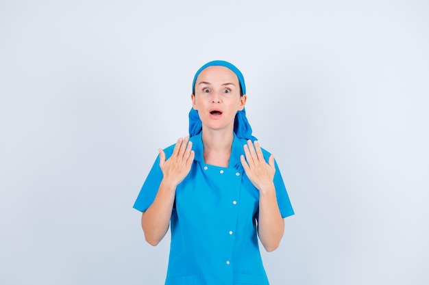 Surprised young nurse is looking at camera by holding up her hands on white background