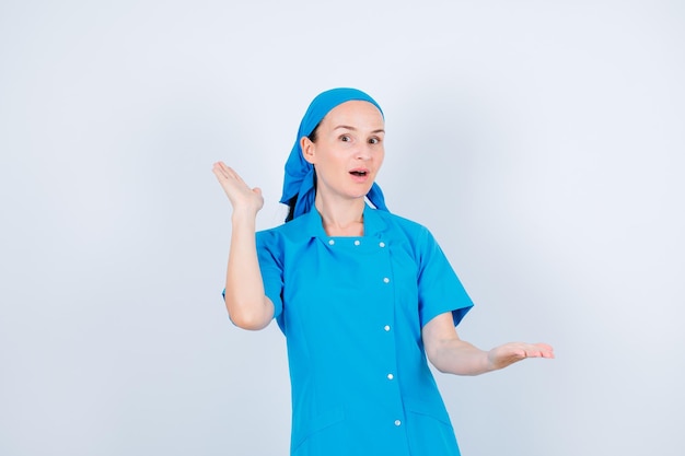 Удивленная молодая медсестра смотрит в камеру, подняв руки на белом фоне