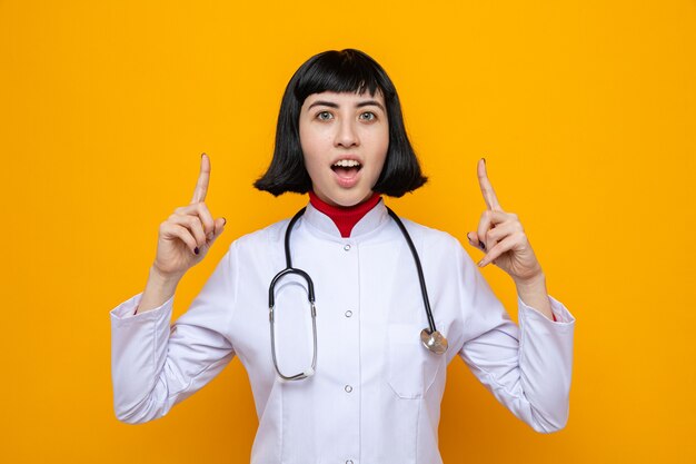 Surprised young pretty caucasian girl in doctor uniform with stethoscope pointing up with two hands