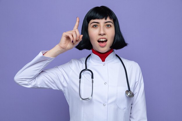 Surprised young pretty caucasian woman in doctor uniform with stethoscope pointing up 