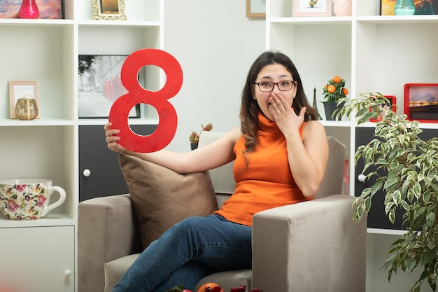 Surprised young pretty girl in optical glasses holding red figure eight and putting hand on her mouth sitting on armchair in living room on march international women's day