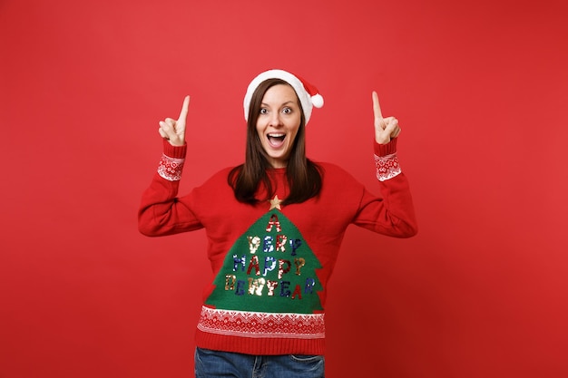 Surprised young Santa girl in Christmas hat keeping mouth wide open and pointing index fingers up isolated on red background. Happy New Year 2019 celebration holiday party concept. Mock up copy space.