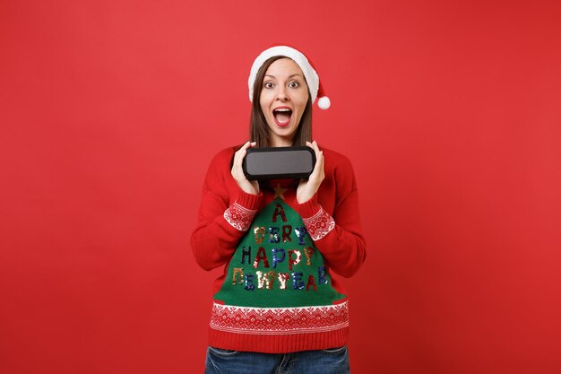 Surprised young Santa girl keeping mouth wide open hold portable wireless bluetooth music speaker isolated on red background. Happy New Year 2019 celebration holiday party concept. Mock up copy space.