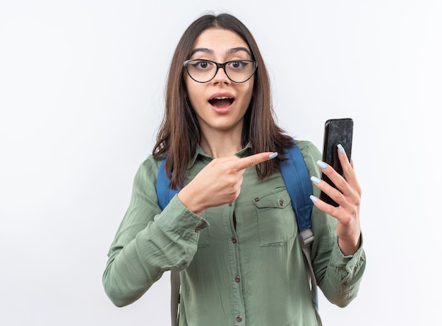 Surprised young school woman wearing glasses with backpack holding and points at phone 