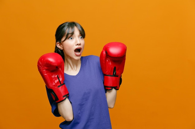 Surprised young sporty woman wearing tshirt and boxing gloves keeping hands in air looking at side isolated on orange background