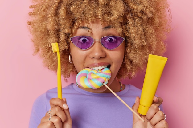 Удивленная молодая женщина кусает разноцветные конфеты, держит зубную щетку и зубную пасту, собирается чистить зубы, носит модные фиолетовые солнцезащитные очки, повседневную футболку, изолированную на розовом фоне. Концепция ухода за полостью рта