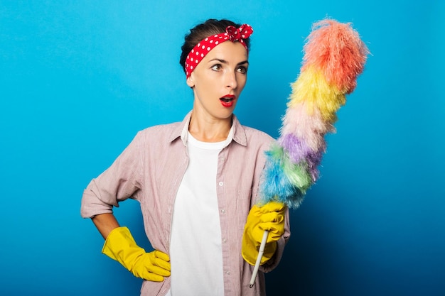 Surprised young woman in gloves holding dust brush on blue background