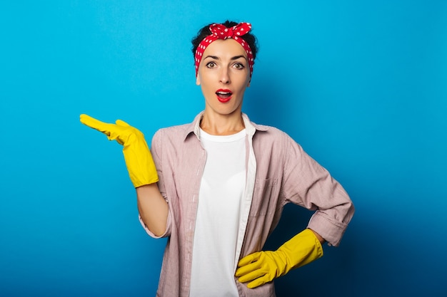 Surprised young woman in gloves holding an invisible object showing thumb up gesture on blue surface