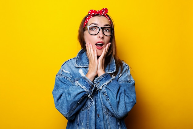 Surprised young woman in hair band, in denim jacket on yellow background.