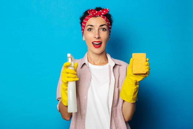 Surprised young woman holding spray and sponge in gloves on blue background.