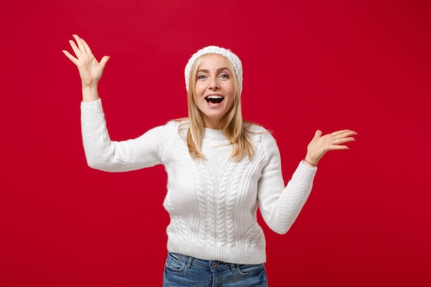 写真 赤い壁の背景のスタジオ ポートレートに分離された白いセーター帽子で驚いた若い女性。健康的なファッション ライフ スタイルの人々 の誠実な感情、寒い季節の概念。コピー スペースをモックアップします。手を広げます。